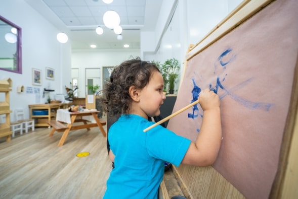 A little girl is drawing on an easel in a classroom.